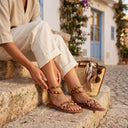 Sandales plates André NOLUNE en cuir camel avec brides croisées et clous dorés, portées en extérieur.