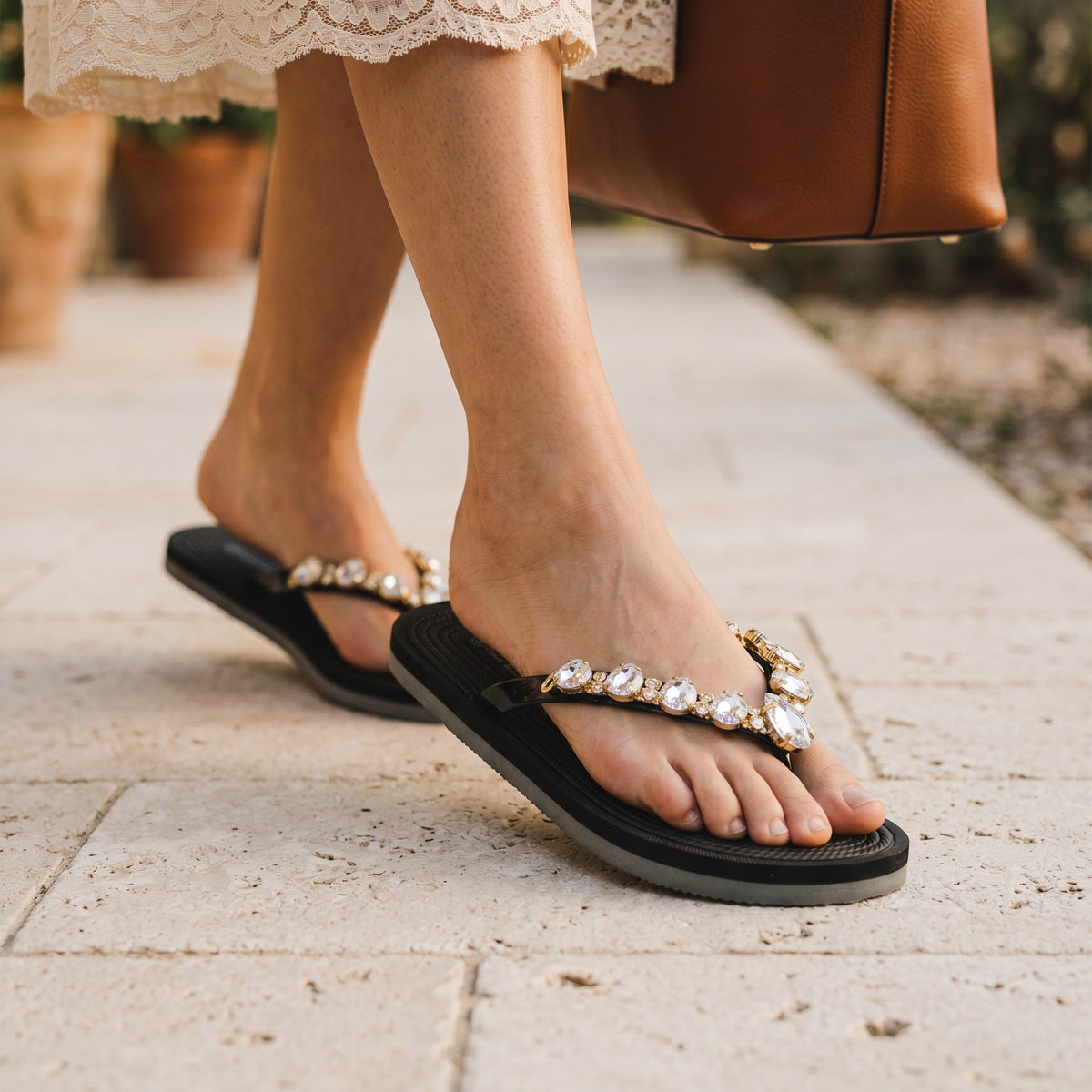 Tongs noires André Orianne ornées de strass brillants portées aux pieds sur un sol en pierre
