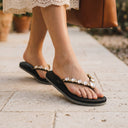 Tongs noires André Orianne ornées de strass brillants portées aux pieds sur un sol en pierre