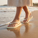 Tongs André Pascale beiges ornées de strass portées sur le sable au bord de l'eau