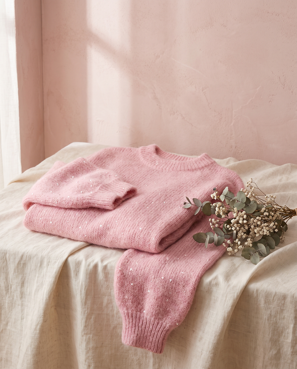 Pull paillettes Solveig rose, posé sur un lin crème, mur rose poudré